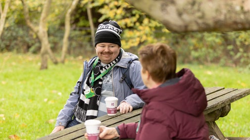 Two people enjoying a hot drink whilst sat a picnic bench.